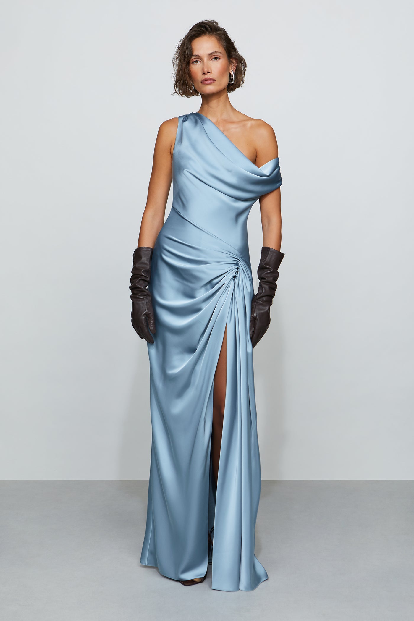 Woman wearing a light blue one-shoulder evening gown with black gloves on a plain background