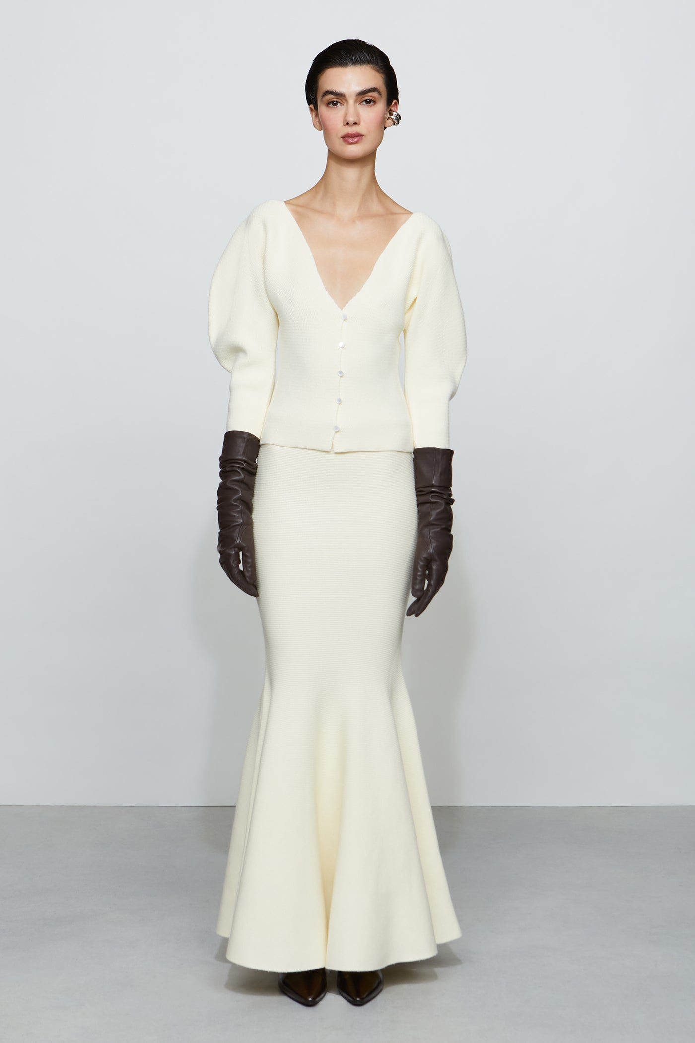 Woman wearing a white outfit with a deep V-neck top and long skirt, paired with dark gloves on a plain background.