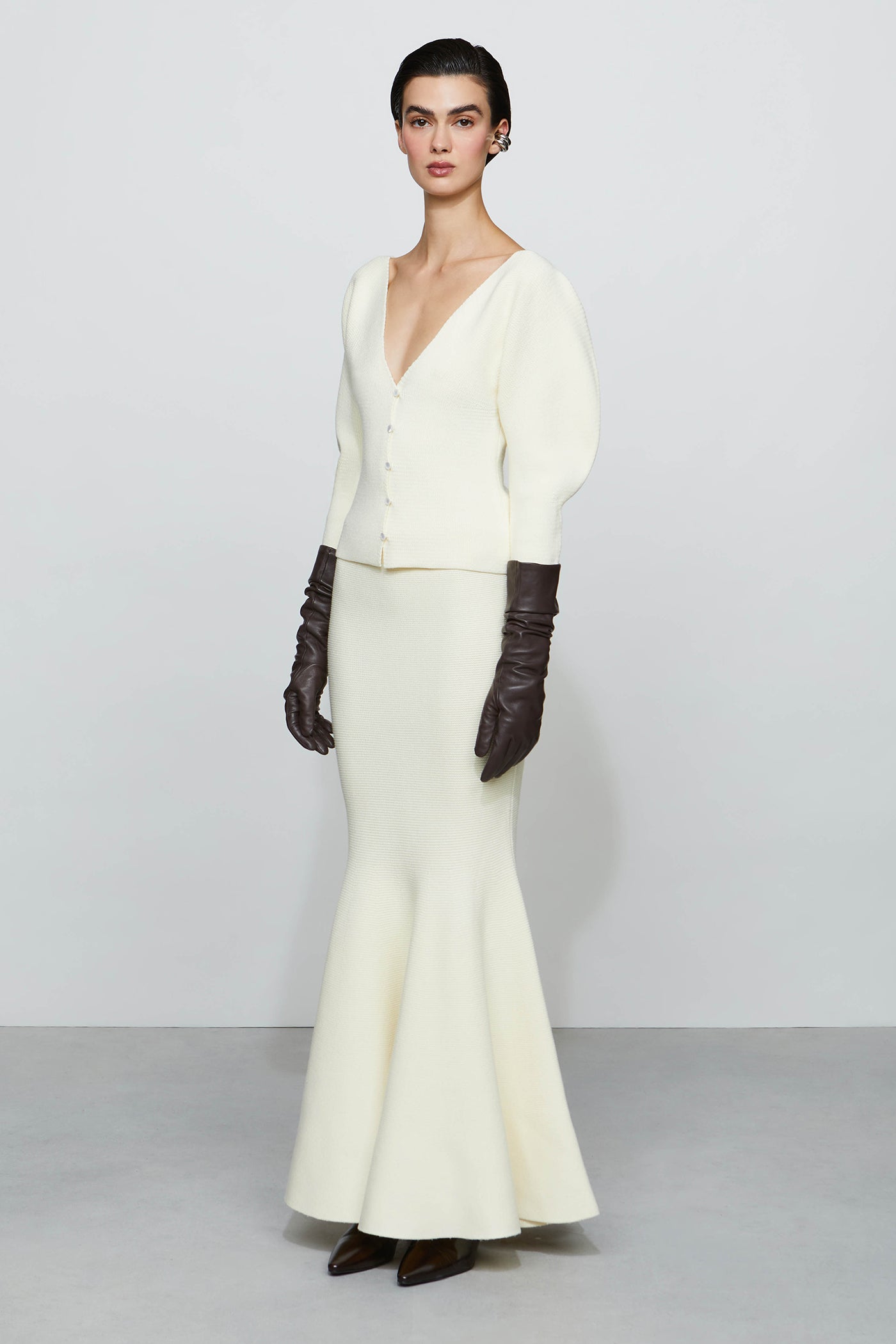 Woman wearing a white outfit with a cardigan and gloves on a plain background