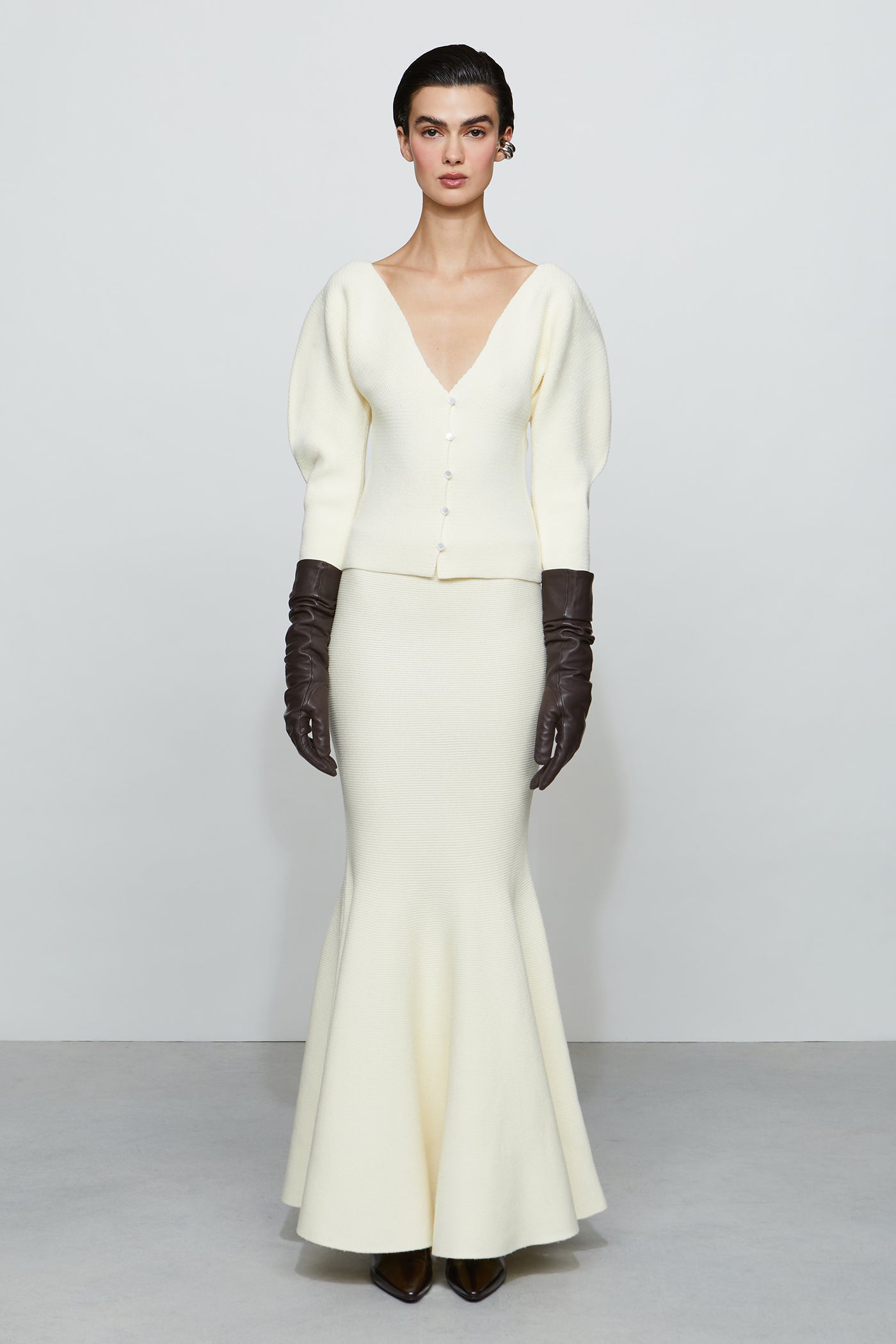 Woman wearing a long white flowing skirt with a fitted shirt, paired with dark gloves on a plain background.