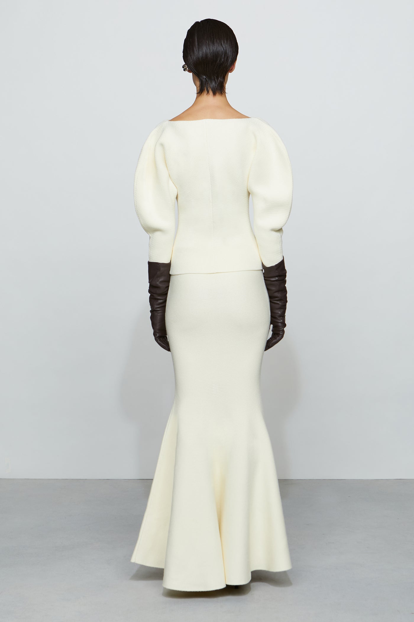 Woman wearing a long white flowing skirt with a fitted shirt, paired with dark gloves on a plain background.