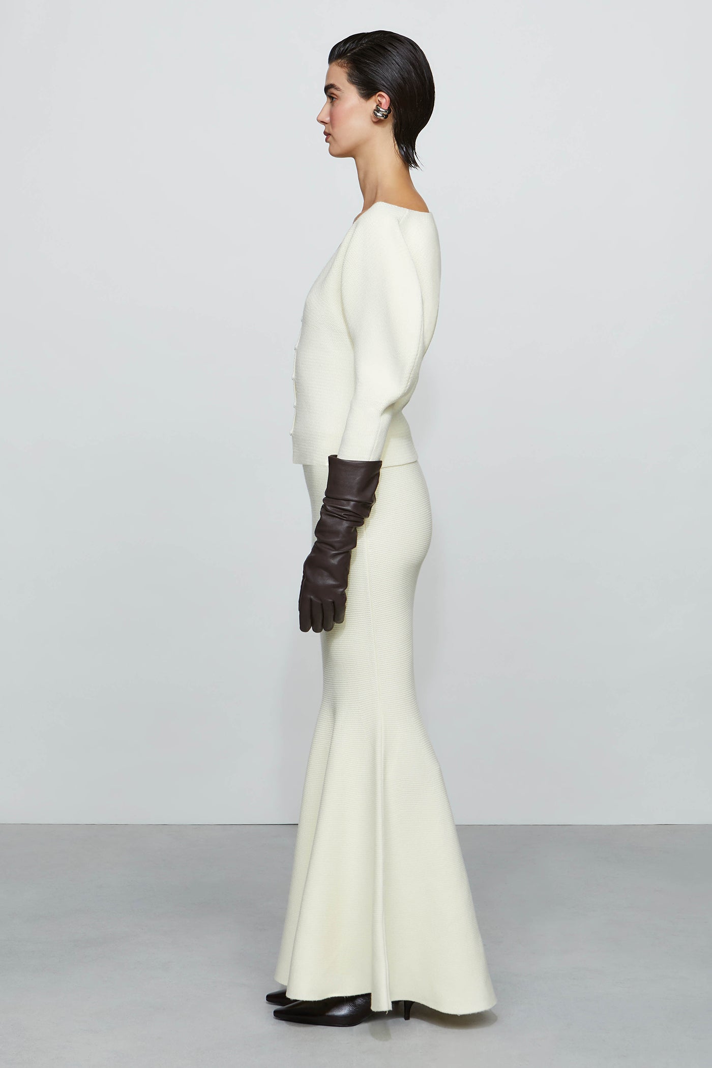 Woman wearing a long white flowing skirt with a fitted shirt, paired with dark gloves on a plain background.