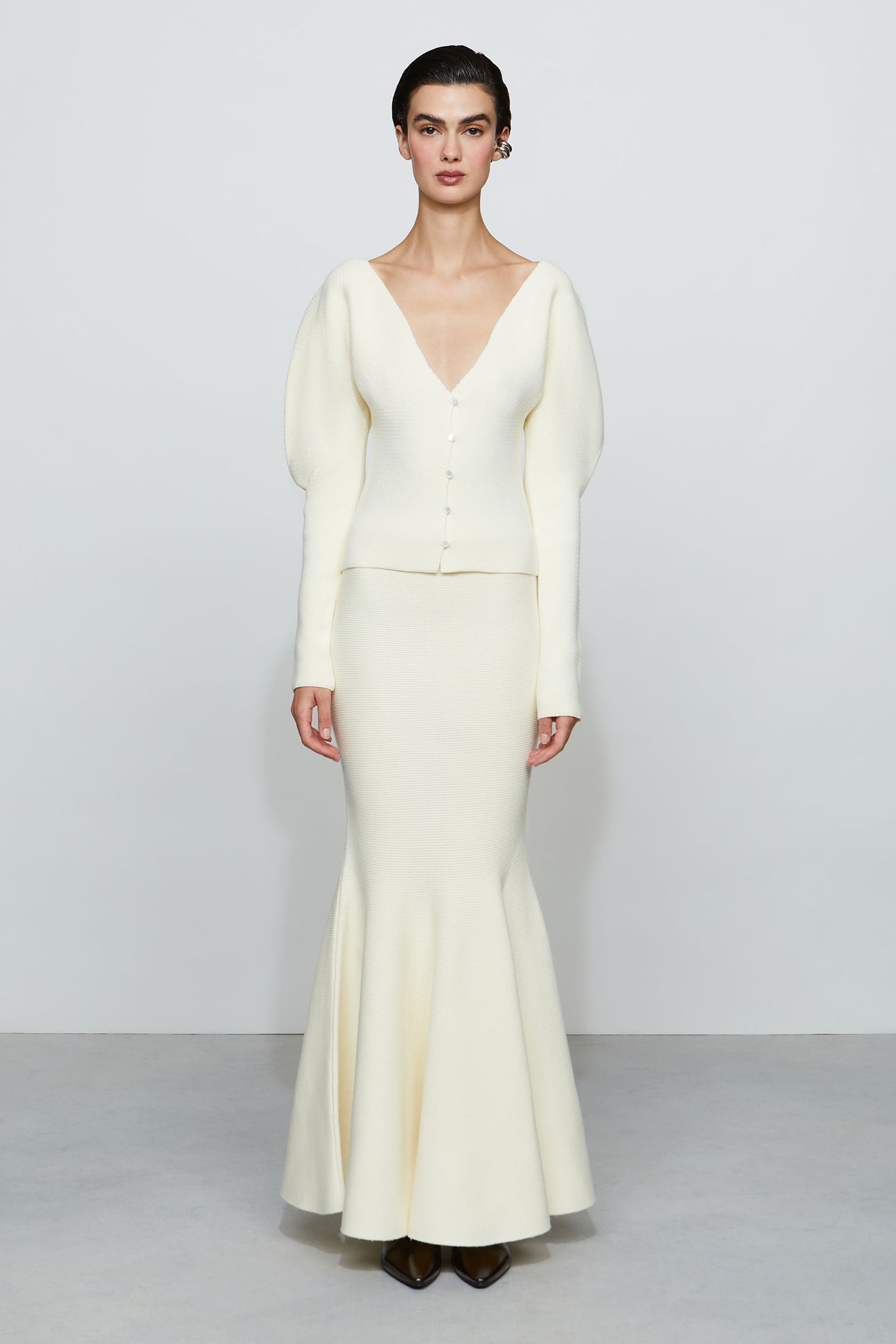 Woman wearing a long, light-colored fitted skirt and top with a v neckline against a plain background.