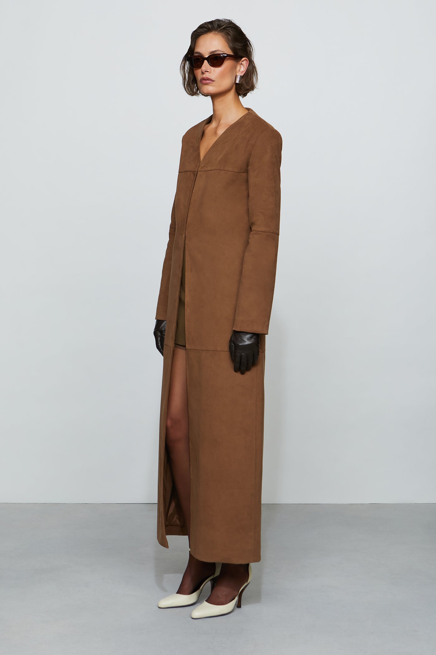 Woman wearing a long brown coat with sunglasses and gloves on a white background